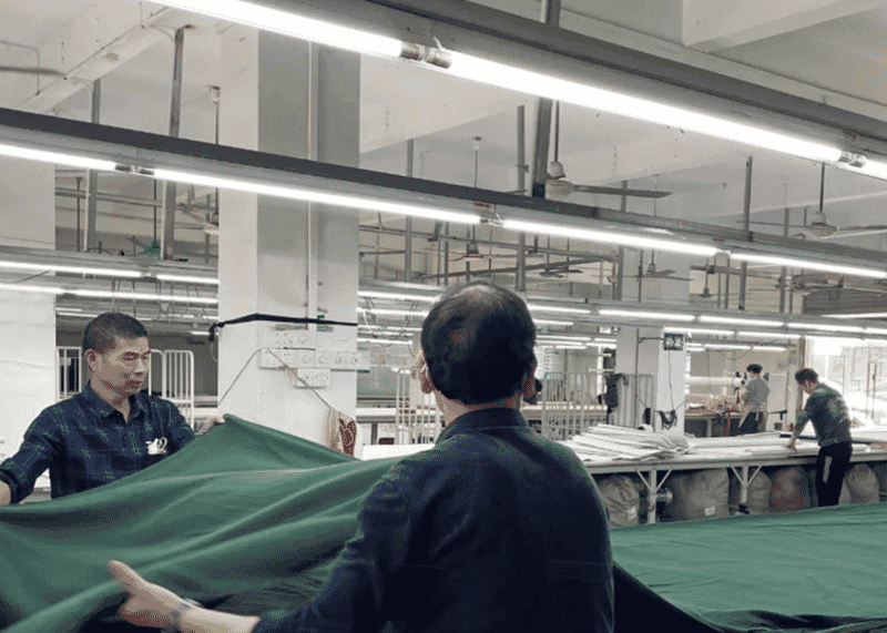 Two workers spreading a length of green fabric on a long cutting table