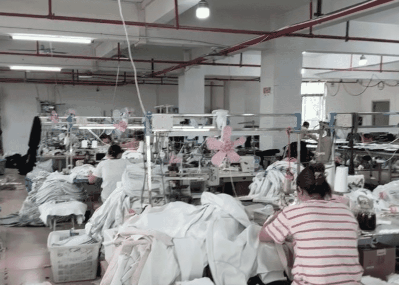 Operators at sewing stations surrounded by piles of light knit panels