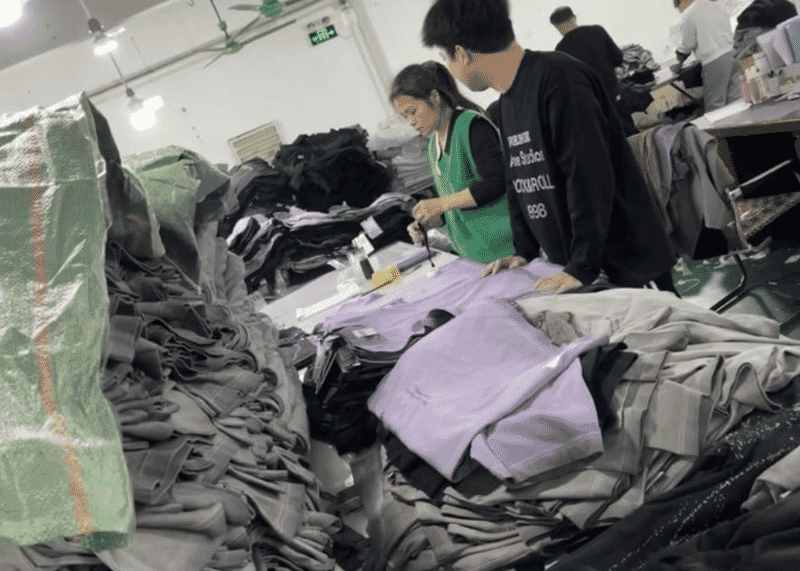 Two workers marking a lilac garment panel on a table piled with grey pieces