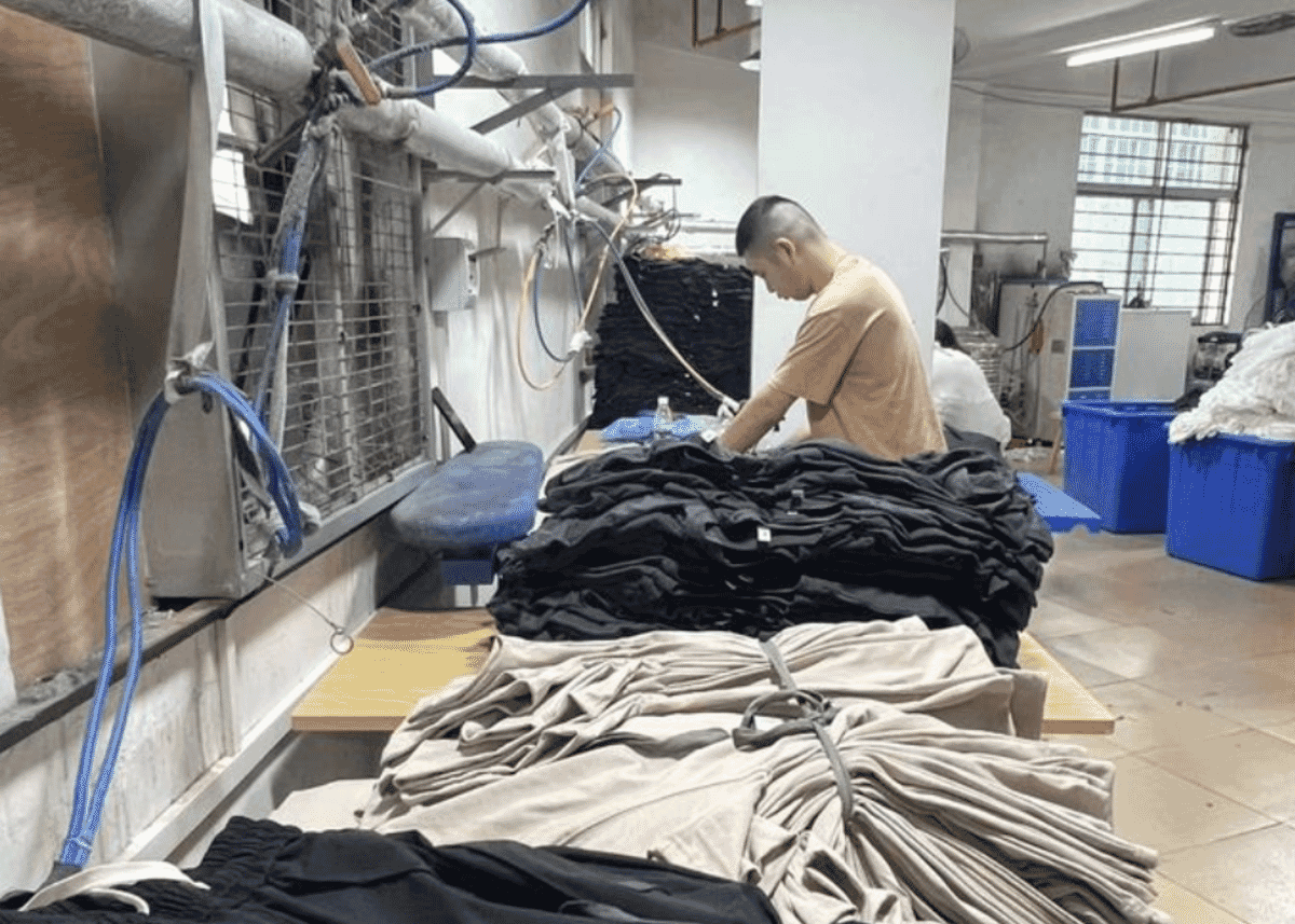 Worker at a pressing station with stacks of black and beige finished garments on the table
