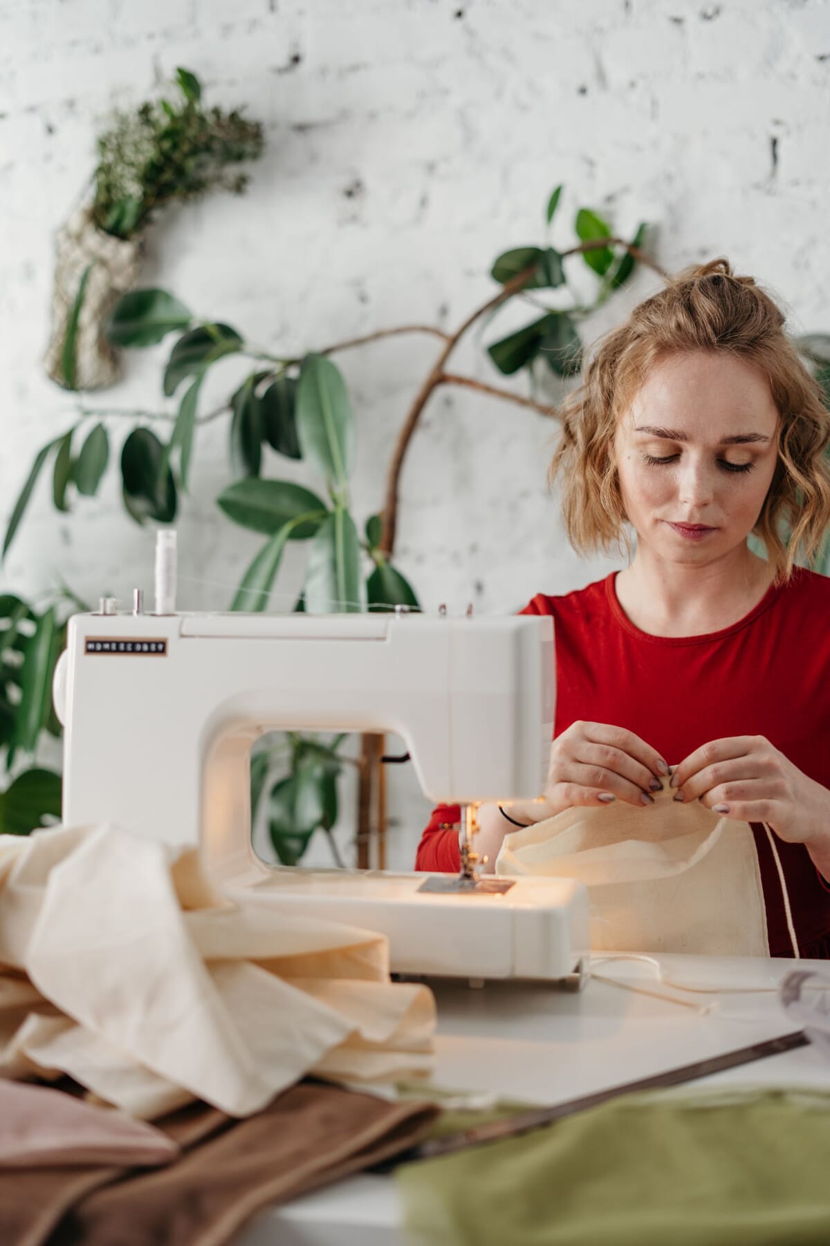 A woman sewing at a domestic sewing machine surrounded by fabric, the sampling stage where a sweatshirt tech pack is tested