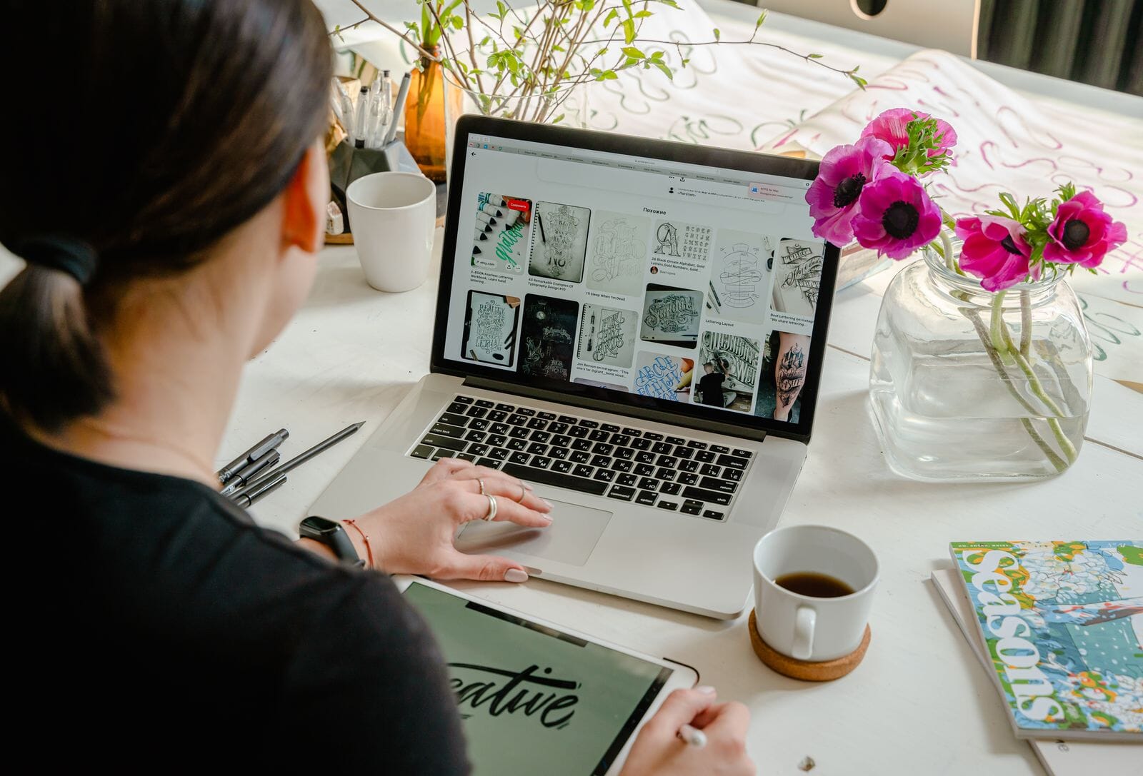 A designer working at a laptop with a graphics tablet and reference material on a bright desk