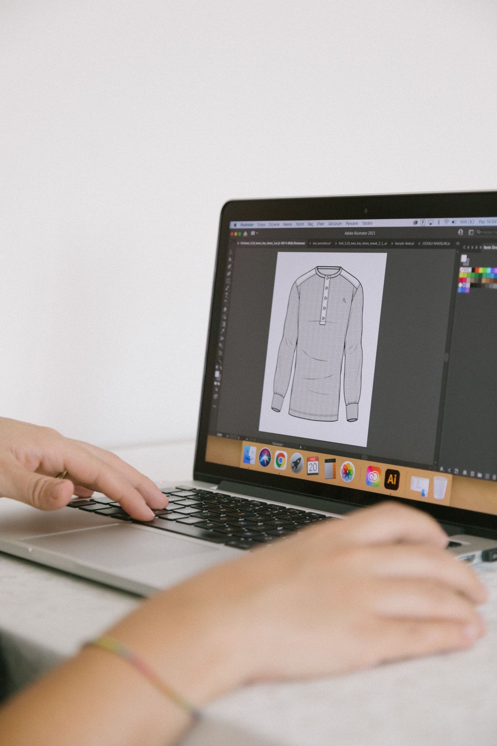 Hands typing on a laptop running Adobe Illustrator with a technical flat sketch of a garment on screen