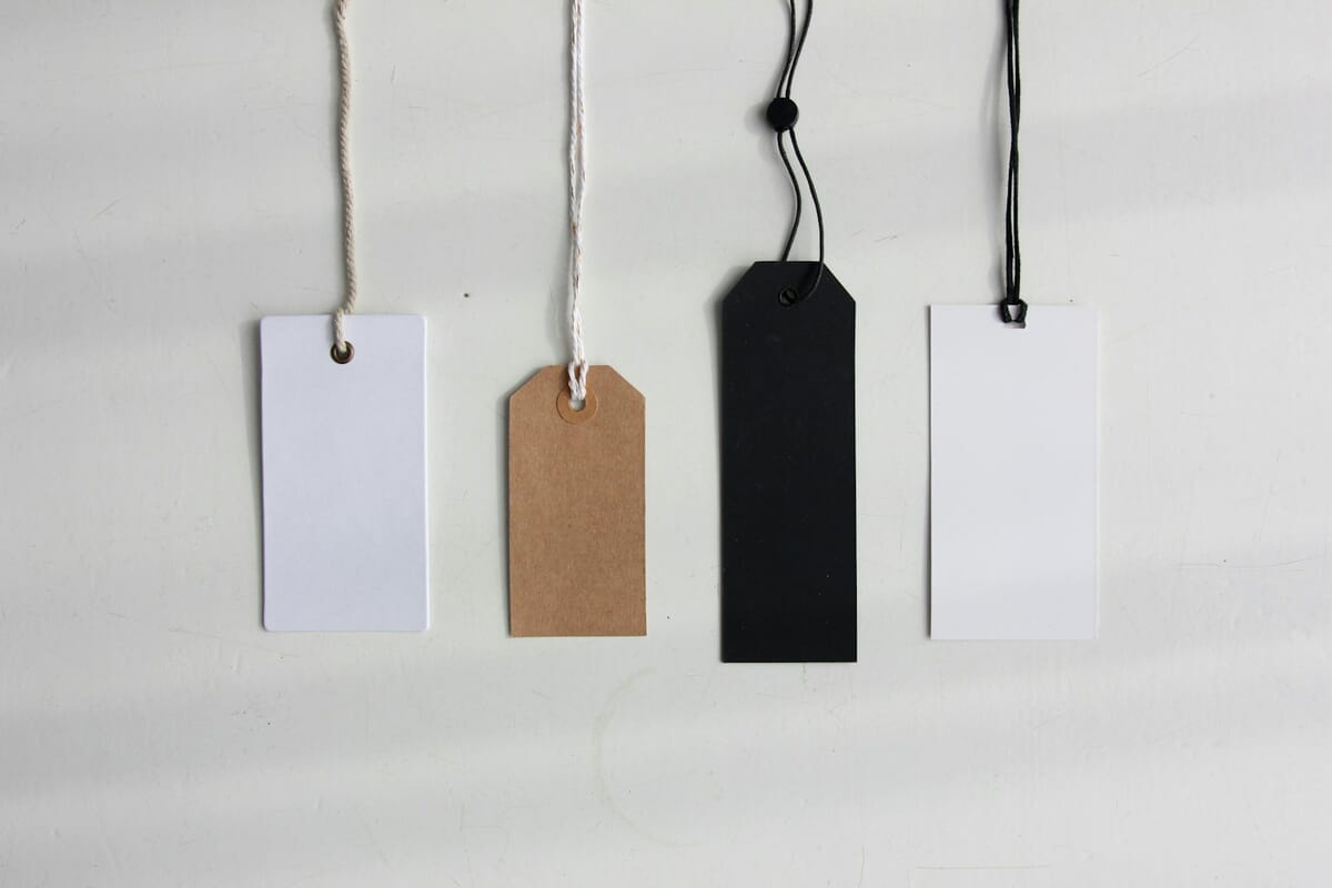 Four blank hang tags in white, kraft brown, black and white hanging on strings against a white wall