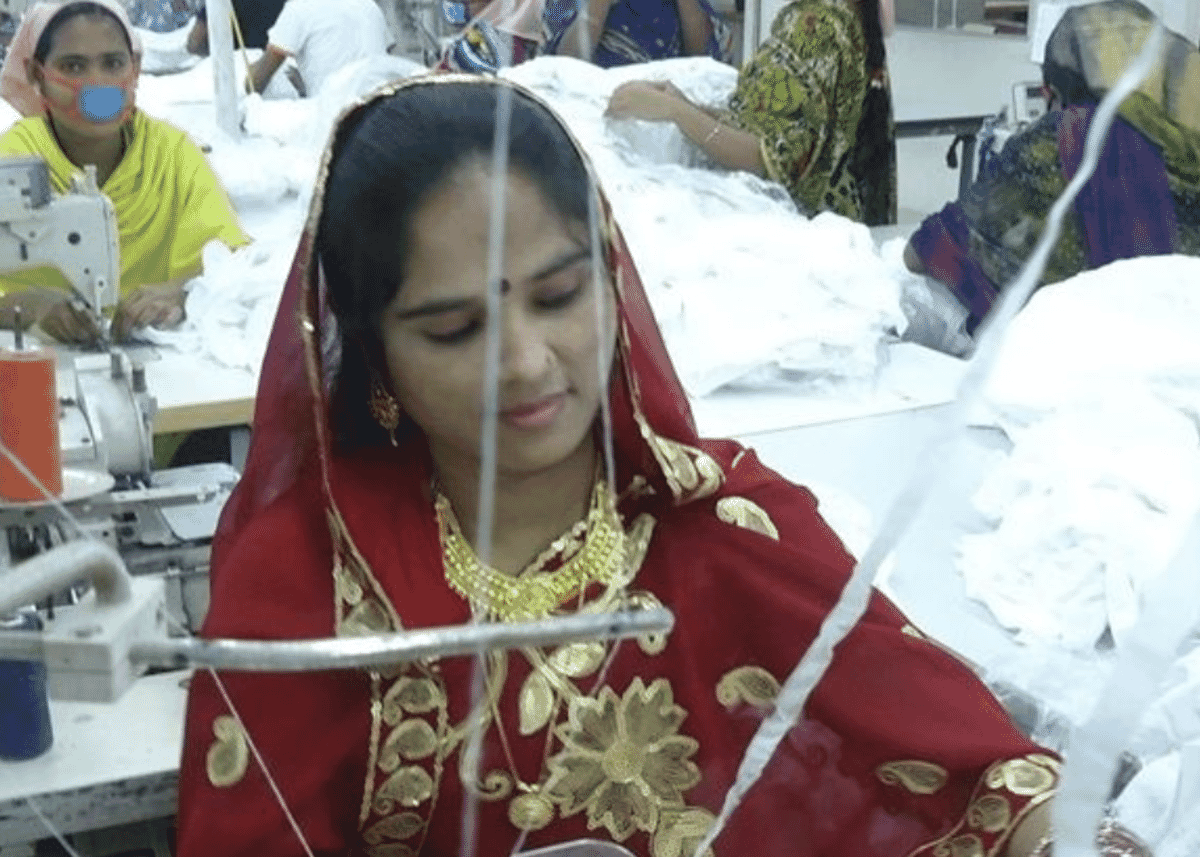 Machinist in a red and gold dupatta sewing white fabric, with colleagues at their machines behind her