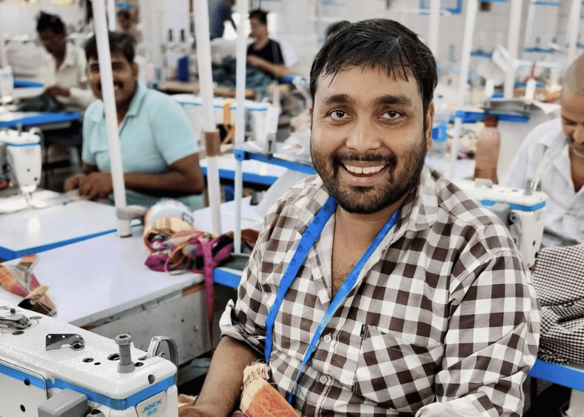 Smiling worker in a checked shirt at a sewing line, with machinists behind him