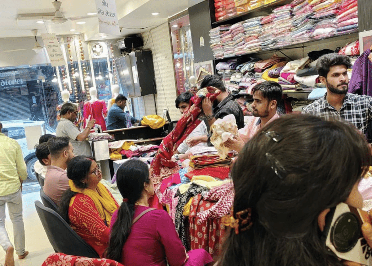 Shoppers and staff looking through printed fabrics and folded salwar suits at a busy clothing shop in India