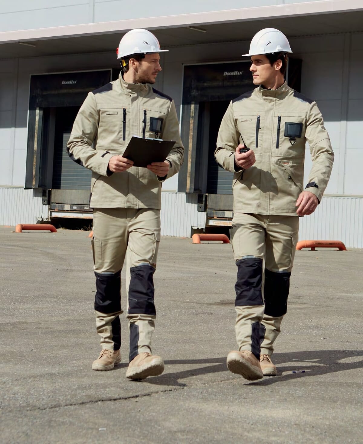 Two workers in beige work jackets and trousers with dark knee panels and white hard hats