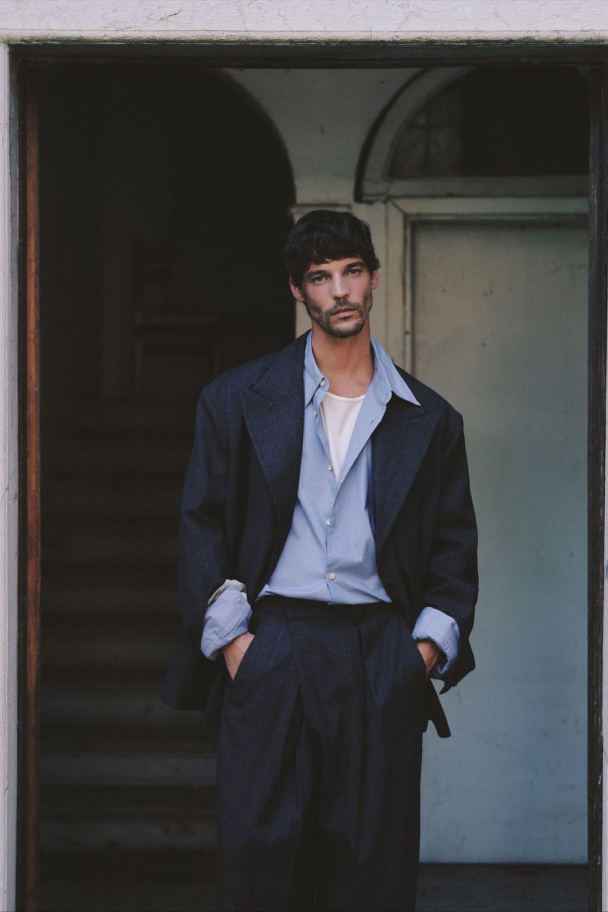Man in a loose navy suit and light blue shirt standing in a doorway