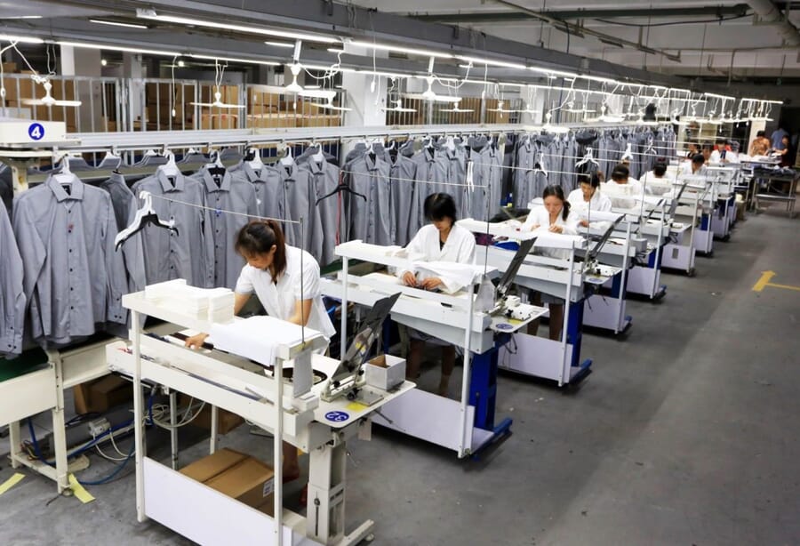 Operators at a line of sewing stations with finished grey shirts on a hanging rail above