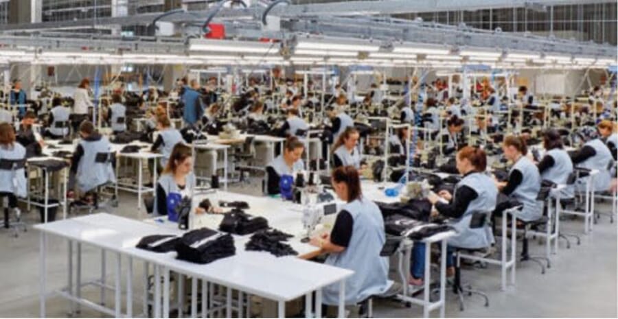 Wide sewing floor with operators in light smocks and dark garments stacked on white tables