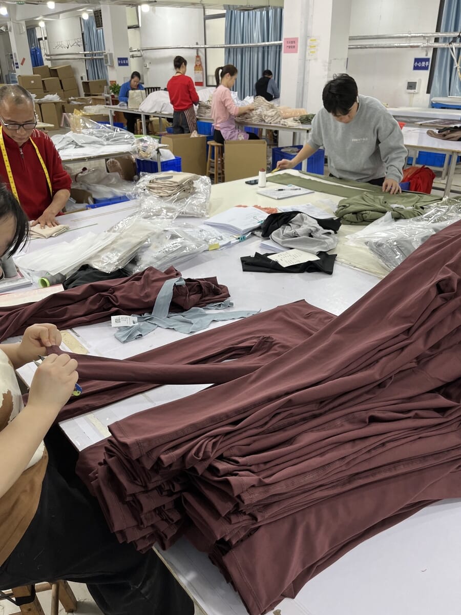 Worker folding brown jersey pieces at a table while others trim behind her