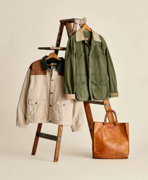 Two utility jackets hung on a wooden ladder with a leather tote