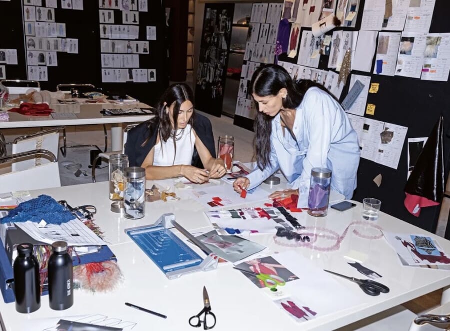 Two designers at a crowded worktable cutting and sketching, with moodboards pinned on the wall behind them