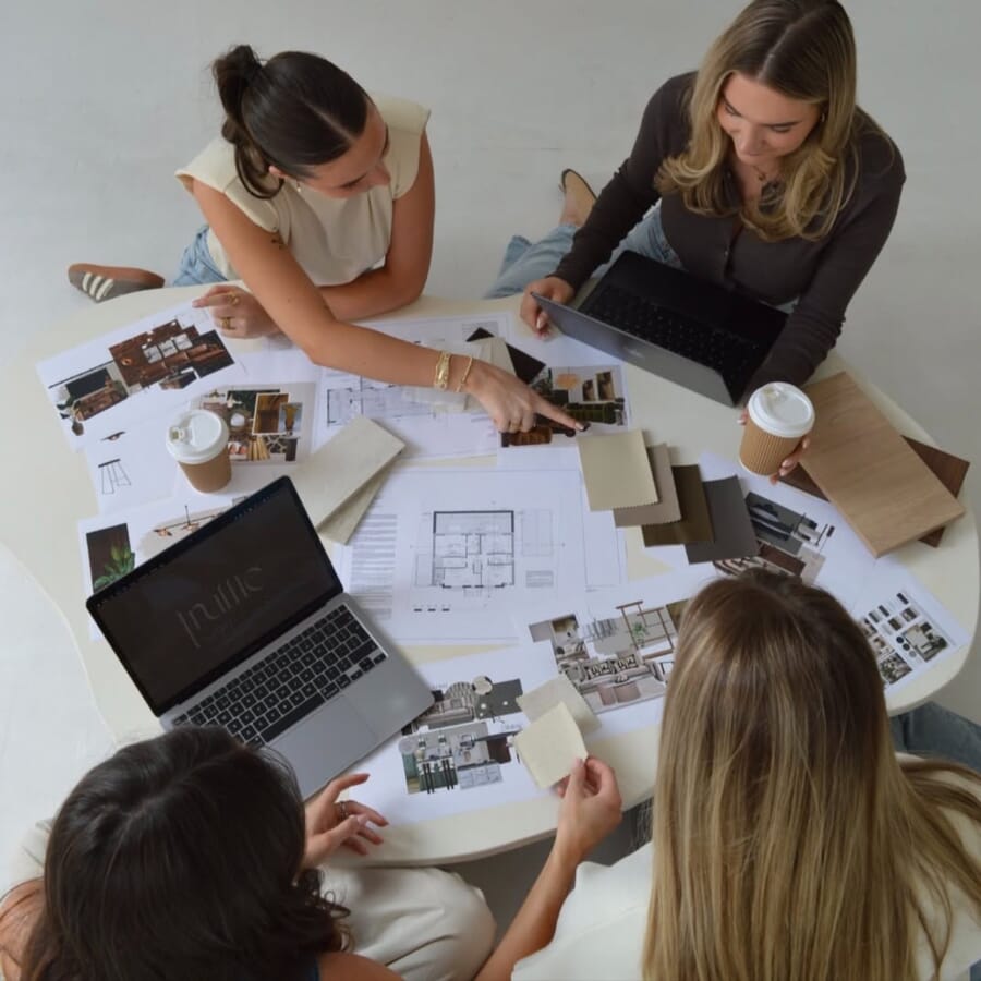 Overhead view of a team around a table with laptops, printed flat sketches, fabric swatches and coffee