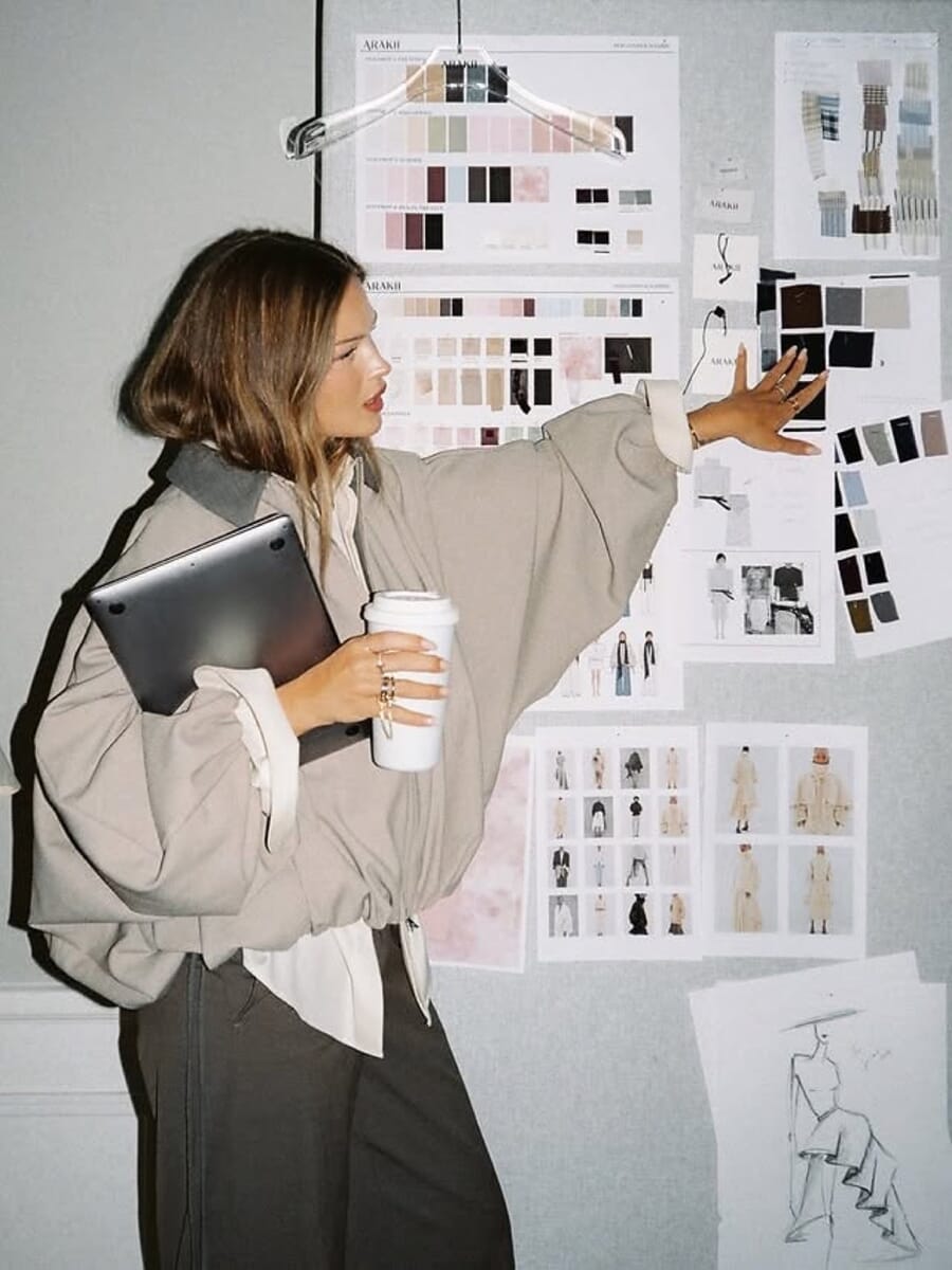 Designer holding a coffee and tablet, pointing at sketches pinned on a wall