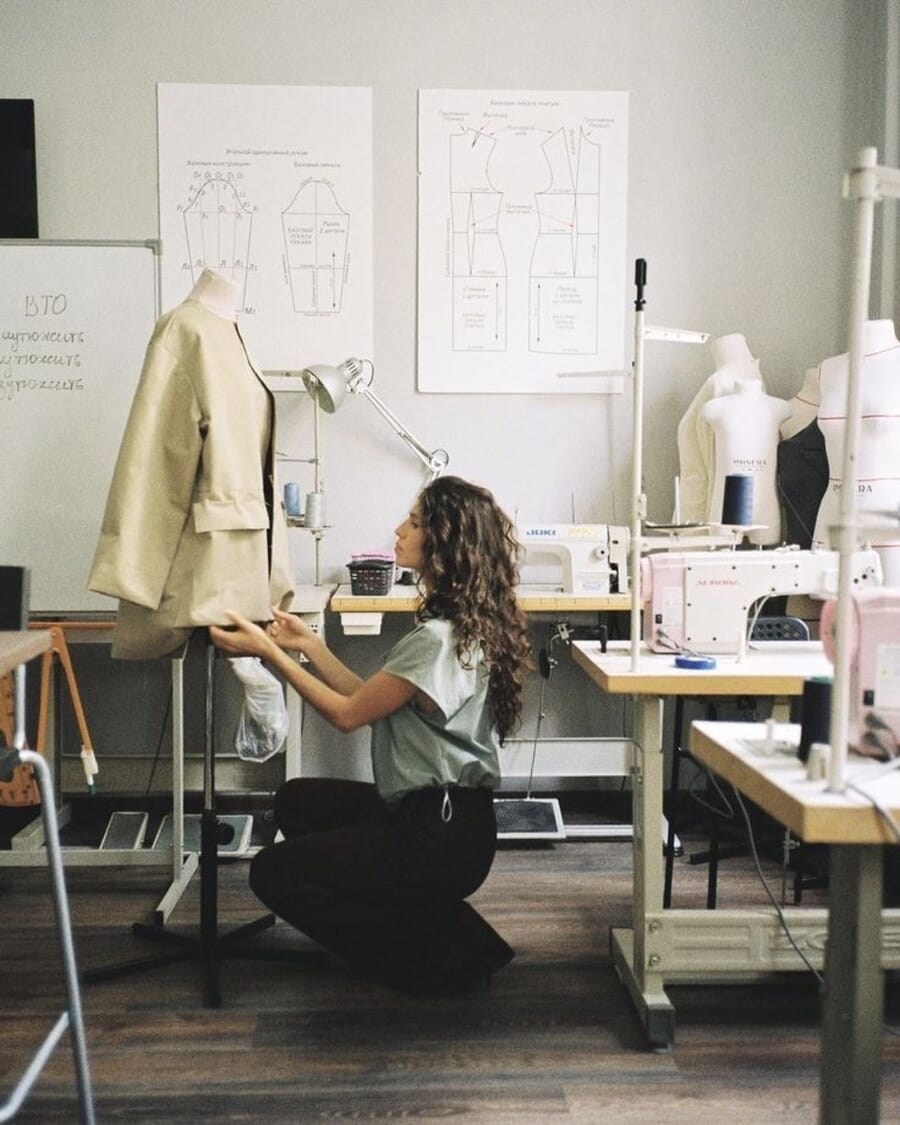 Designer kneeling to adjust a cream jacket on a dress form in a sampling room