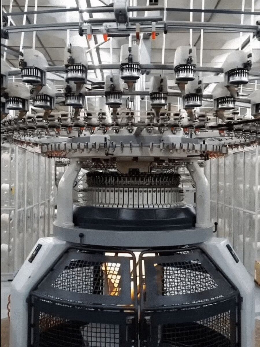 Circular knitting machine with yarn feeders around the top, knitting a tube of jersey