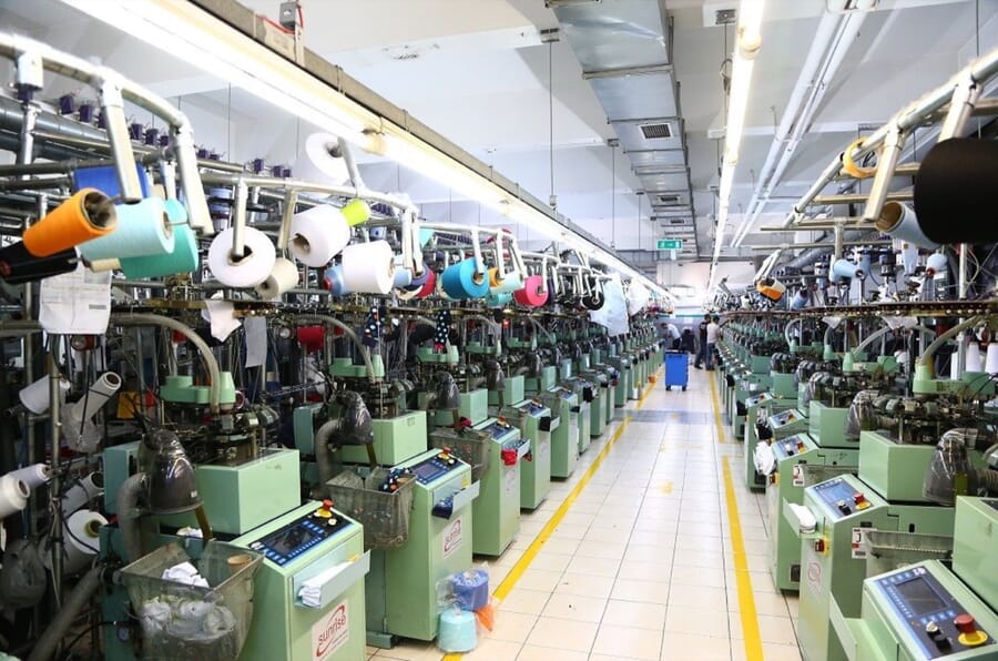 Long aisle of knitting machines with colourful yarn cones above them