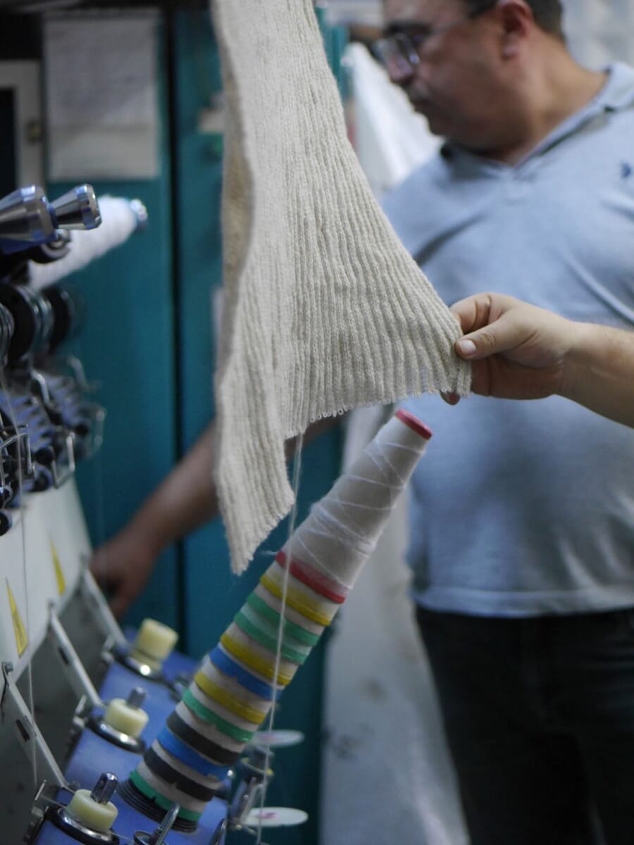 Technician holding up a cream knitted panel beside yarn cones on the machine