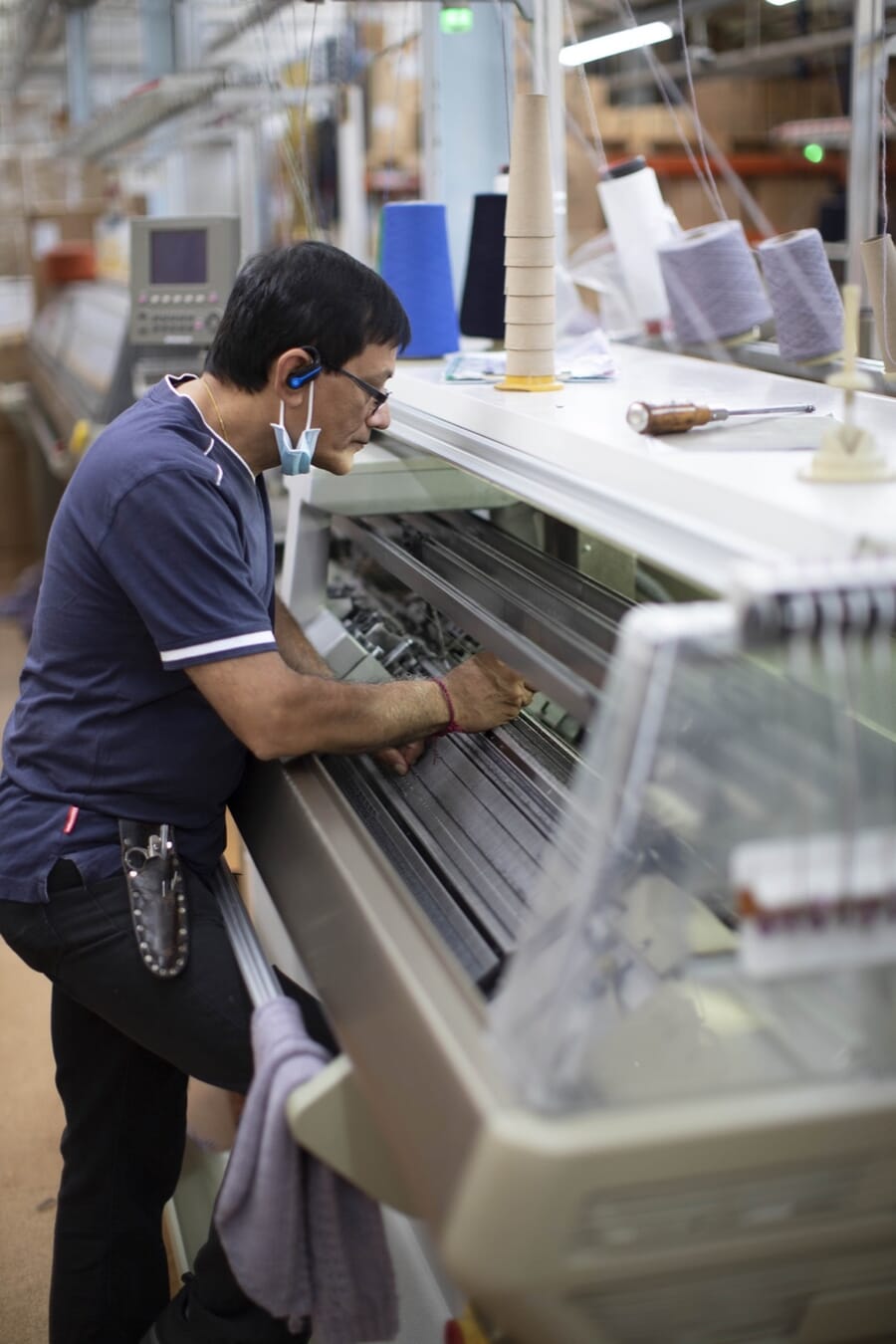 Operator working at a flat knitting machine with yarn cones above