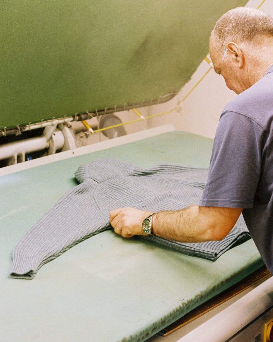 Worker smoothing a grey knitted sweater flat on a steam press table