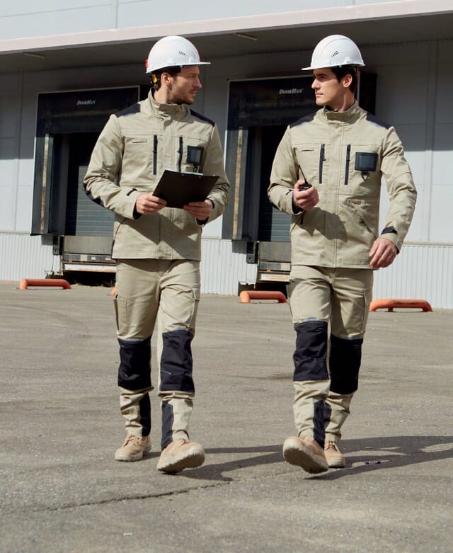 Two engineers in khaki and navy work jackets and trousers with white hard hats, walking past loading docks