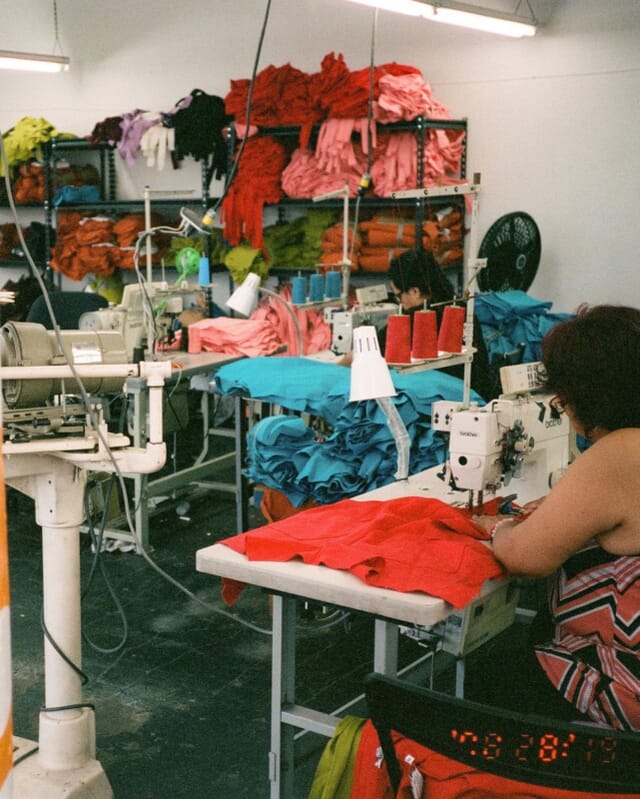 Operator sewing red garments with stacks of colourful cut pieces on the shelves behind