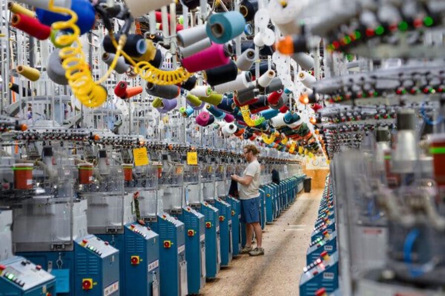 Long rows of sock knitting machines with coloured yarn cones overhead and an operator checking a machine