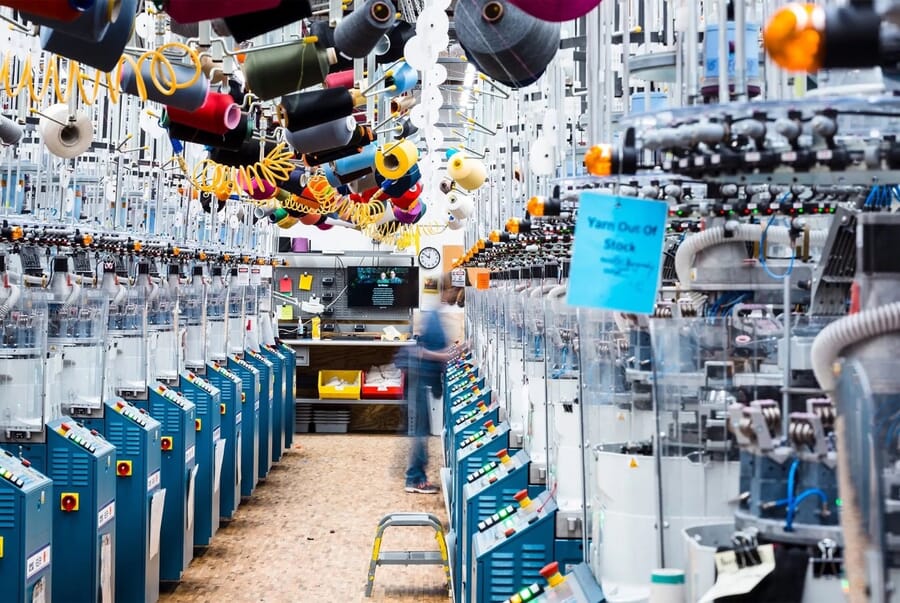 Rows of knitting machines with yarn cones hanging above the aisle