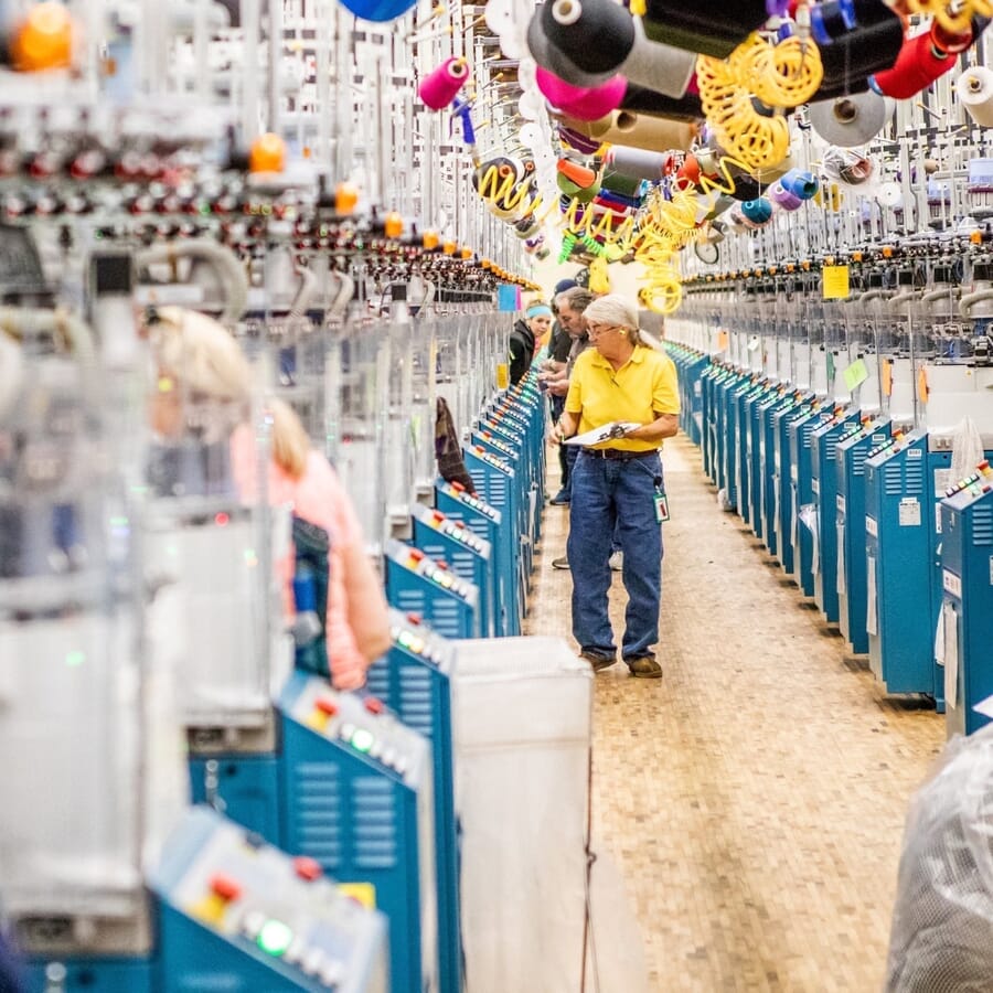 Operator walking the aisle of a sock knitting floor with yarn cones overhead