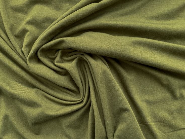 Rolls of soft, drapey cellulosic fabric in warm neutral tones stacked on a wooden shelf at a Portuguese mill, showing the smooth surface characteristic of modal.