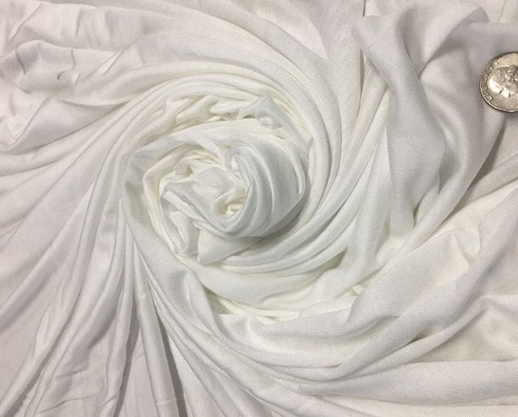 Rolls of soft, drapey off-white lyocell fabric stacked on a wooden shelf at a Portuguese textile mill, showing the smooth surface characteristic of Tencel.