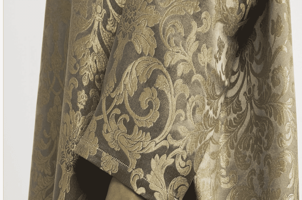 Detailed close-up of a woven jacquard fabric surface with raised botanical motifs in cream and gold tones, showing yarn floats characteristic of loom-woven figured cloth.
