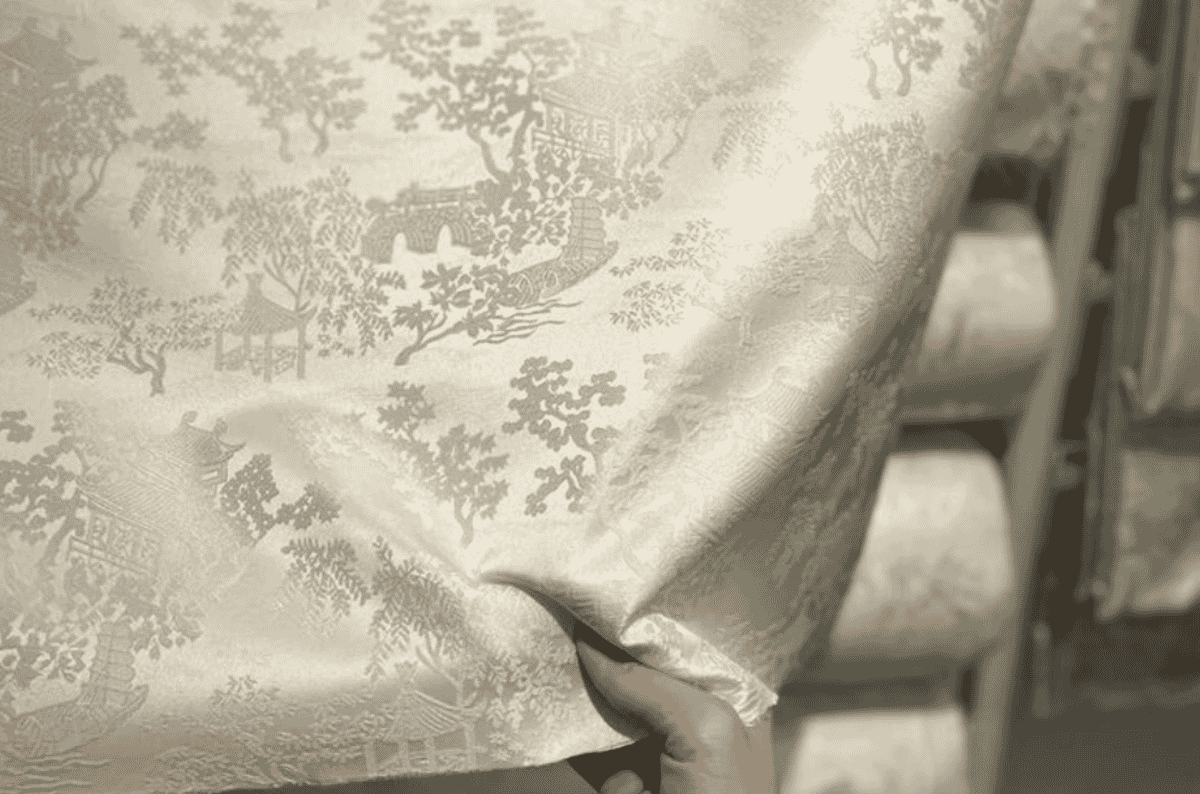 A hand holding ivory scenic jacquard cloth woven with trees, pavilions and figures in a single tone, a chinoiserie-style figured weave.