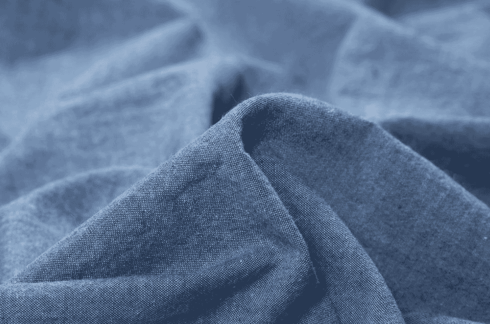 Folded light-blue chambray shirt on a wooden table showing the soft heathered plain-weave surface produced by an indigo warp and white weft.