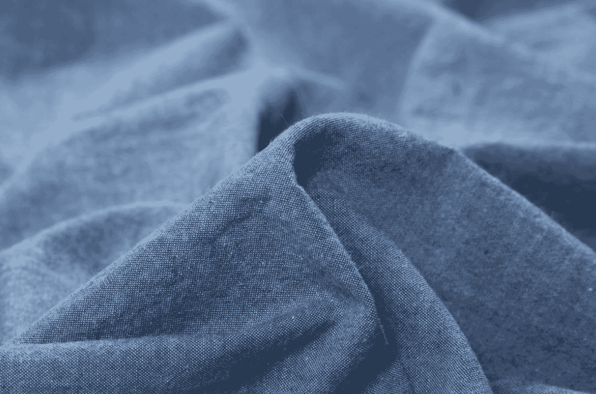 Folded light-blue chambray shirt on a wooden table showing the soft heathered plain-weave surface produced by an indigo warp and white weft.