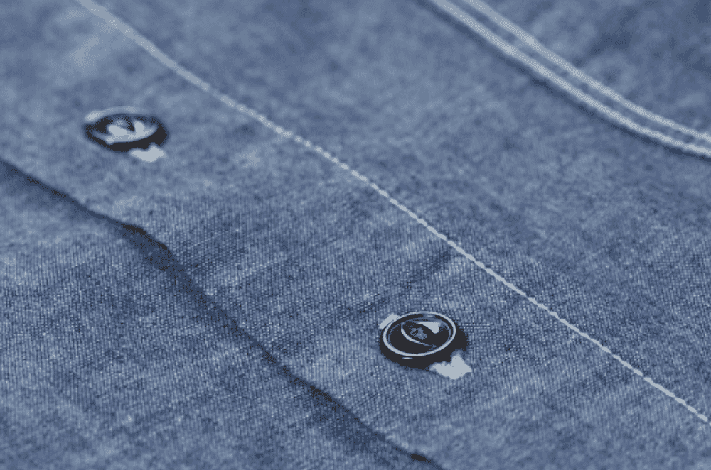 A rail of finished light blue chambray shirts inside a Portuguese factory showing the soft heathered surface and casual button-up construction typical of shirt-weight chambray.