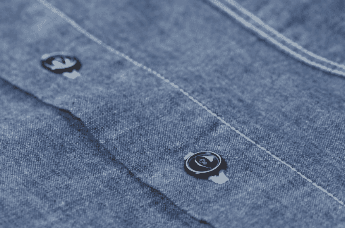 Macro close-up of a darker blue chambray placket with two buttons and a line of topstitching, showing the salt-and-pepper mix of dyed and undyed yarns.
