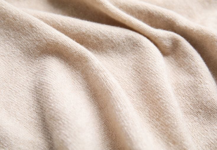 Soft folded cashmere knitwear in neutral cream and beige tones stacked on a wooden surface, showing the fine fibre texture typical of luxury knitted garments.