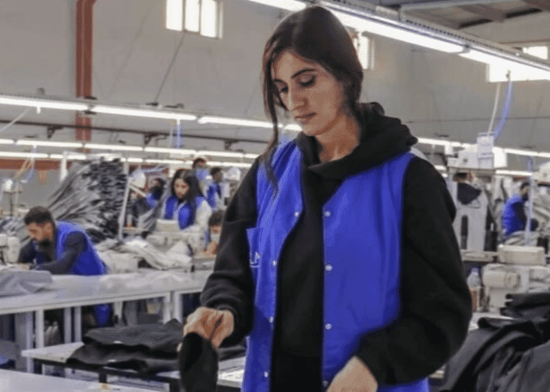 Operator in a work vest handling a garment on a long, brightly lit sewing floor