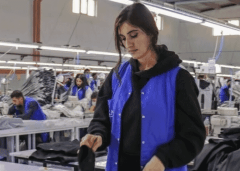 Operator in a work vest handling a garment on a long, brightly lit sewing floor