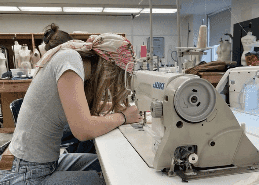 A machinist sewing at an industrial lockstitch machine in a sample room, dress stands behind her